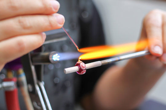 pictured: a person adding details to a red glass bead over a flame. 